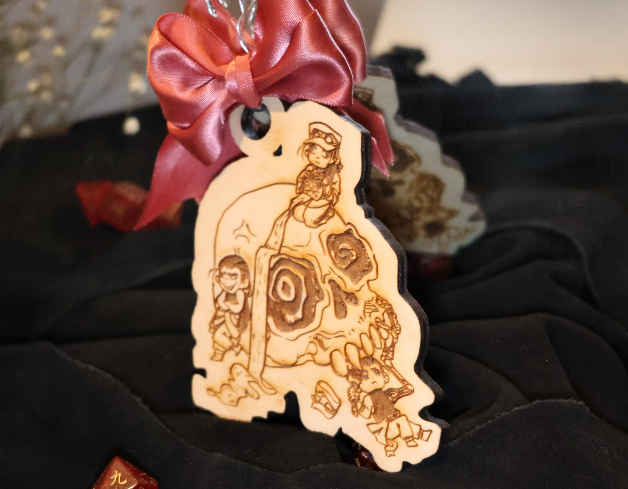 Double-sided ornament with the same laser engraving of art on both sides. The scene depicts a prominent stylized skull with three small people interacting with it. The people all appear to be the same with minor differences. The person on the top of the skull is sitting in seiza (kneeling) while knitting an absurdly long scarf that trails down the oversized skull onto the floor. The distinguishing feature of them is they are wearing a hat. The figure to the left of the skull is leaning on the skull arms crossed. They look devilish with a smile of sharp teeth. They are not wearing a hat. The last figure is holding up the skull with an oversized skeletal hand and arm and in their other hand they have a wrench, as though they are working on the underbody of a mechanical object. Their hat is on the ground next to them. This ornament is cutting around this scene making an irregular shape with a ribbon bow is tied to the top. The ribbon is reddish pink.