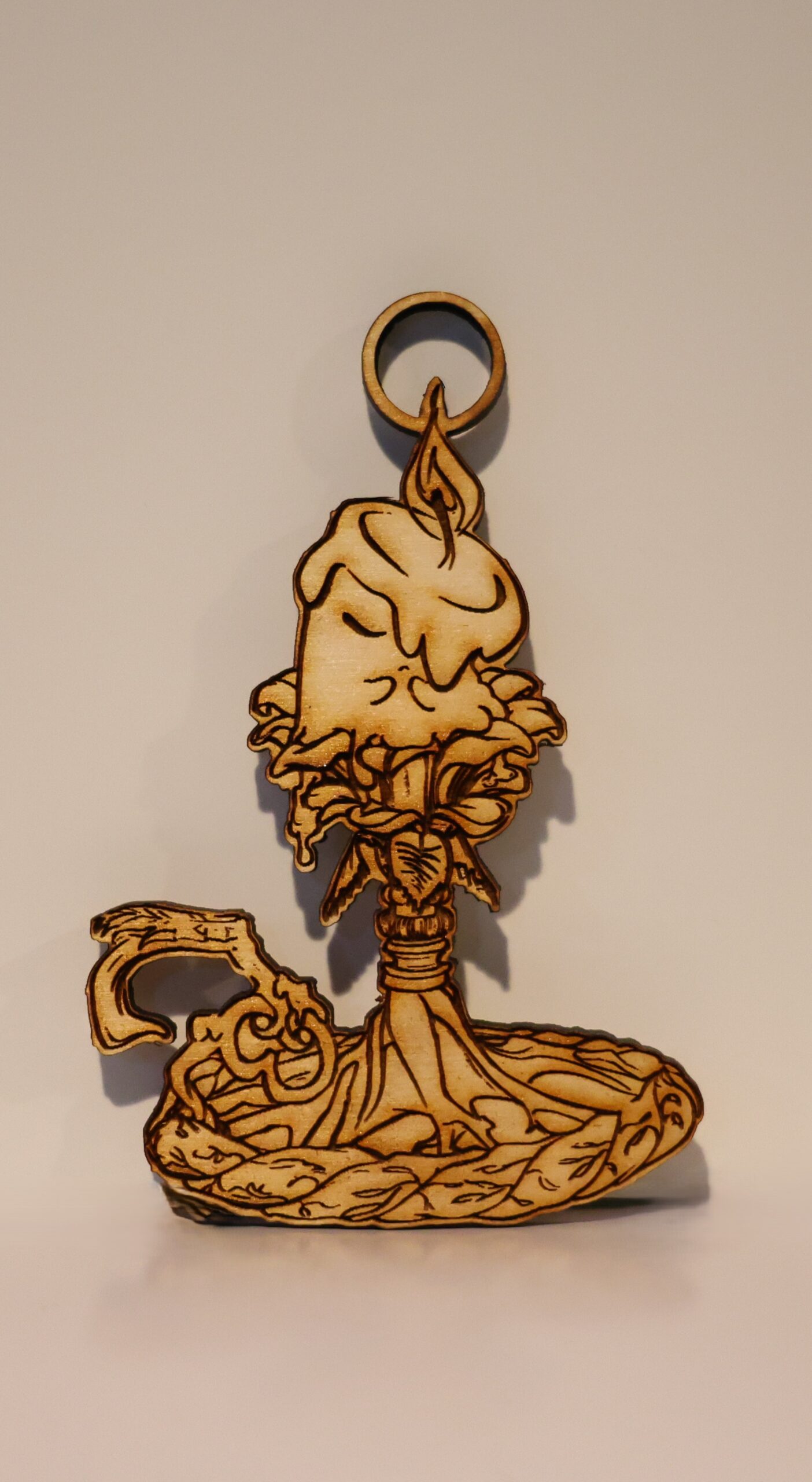 An ornament of a living candle with small arms and two eyes (one hidden under wax dripping down their face) sitting on an ornate candle stick holder that is floral themed. It is a double-sided laser engraved piece of 6 mm wood. The candle is meant to be a stylized litwick from Pokemon.